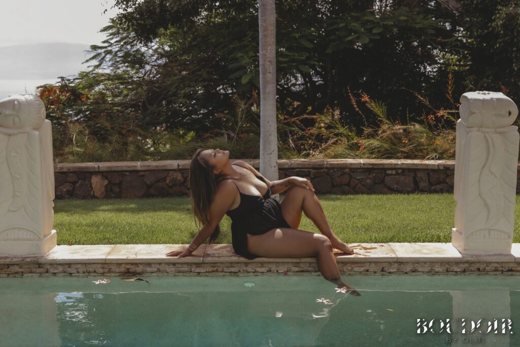 Woman sitting along a pool, one foot in the water with her head thrown back. There are statues on each side of her and foliage in the background behind her.