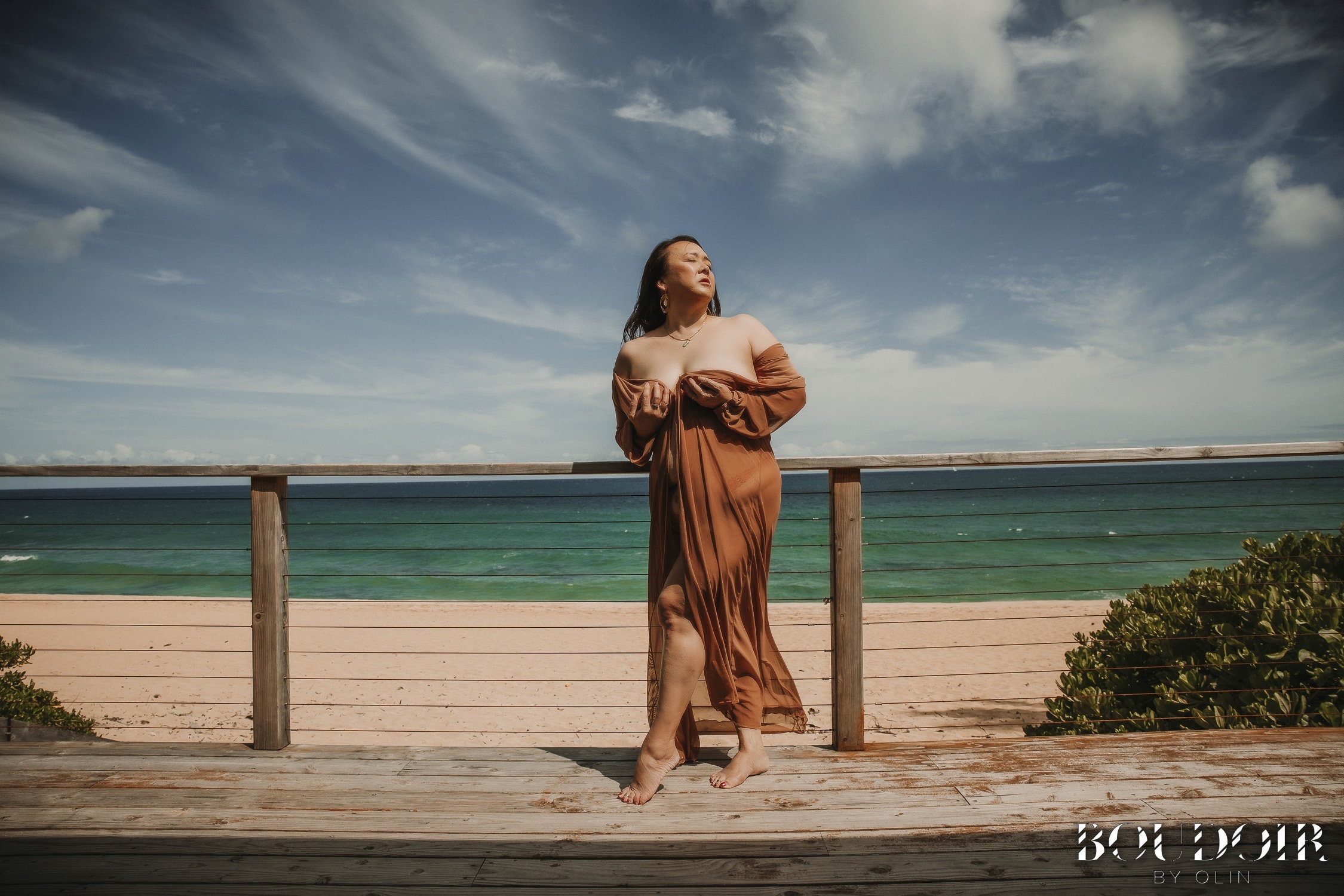 woman in orange dress is standing on a deck with the beach and ocean behind her. it is a bright and beautiful day.